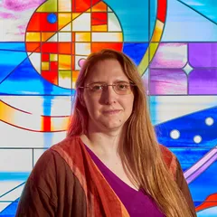 A woman with very long dark blonde honey-colored hair stands in front of a stained glass window showing stars and planets. She wears rimless glasses and a brown and orange Viking tunic coat over a purple shirt. Her right arm is extended, supporting her with a cane.