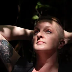 Sarah Gailey, a white person with auburn hair that is long on top and shaved on the sides, looking up with their hands behind their head. Dappled sun and shadow falls across their face, chest, and arms.