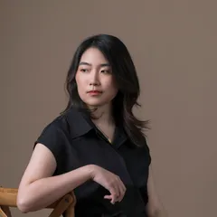 A Chinese person sits on a wooden chair against a brown background, looking to the left. They are wearing a black top and beige trousers.